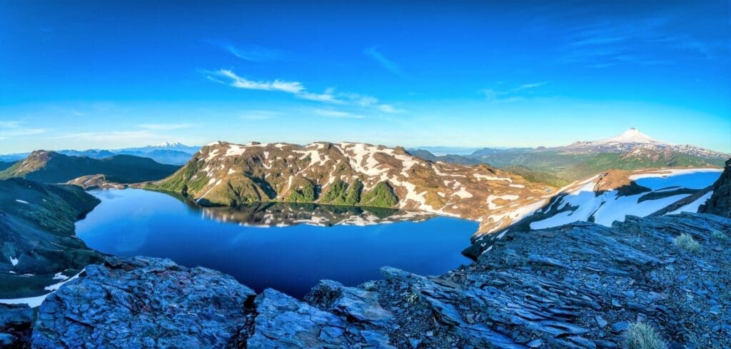 clear lake surrounded by snow-dusted mountains and rocky cliffs on a bright day with volcanoes in the background in South America