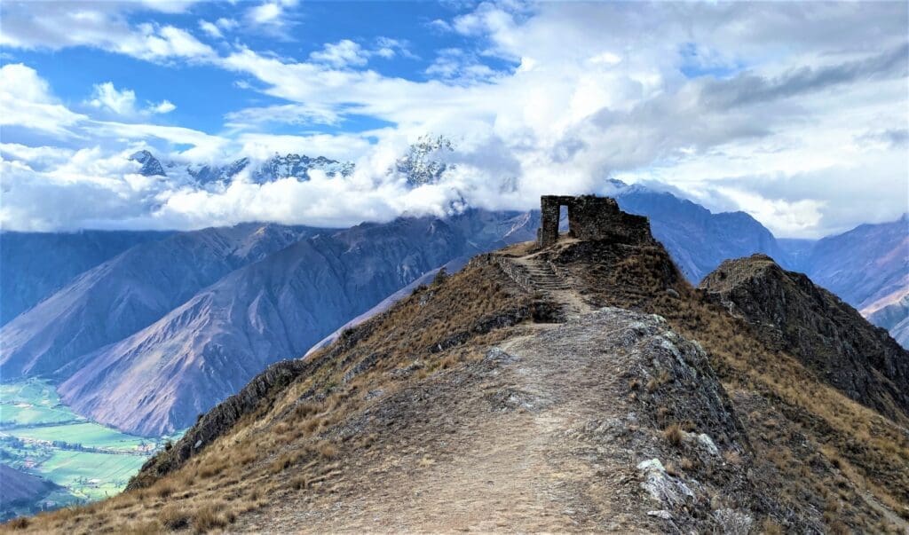 Rocky dirt path leading up to the Sun Gate stone archway overlooking a valley and majestic mountains peeking through clouds on the Inca Trail Express route in Peru