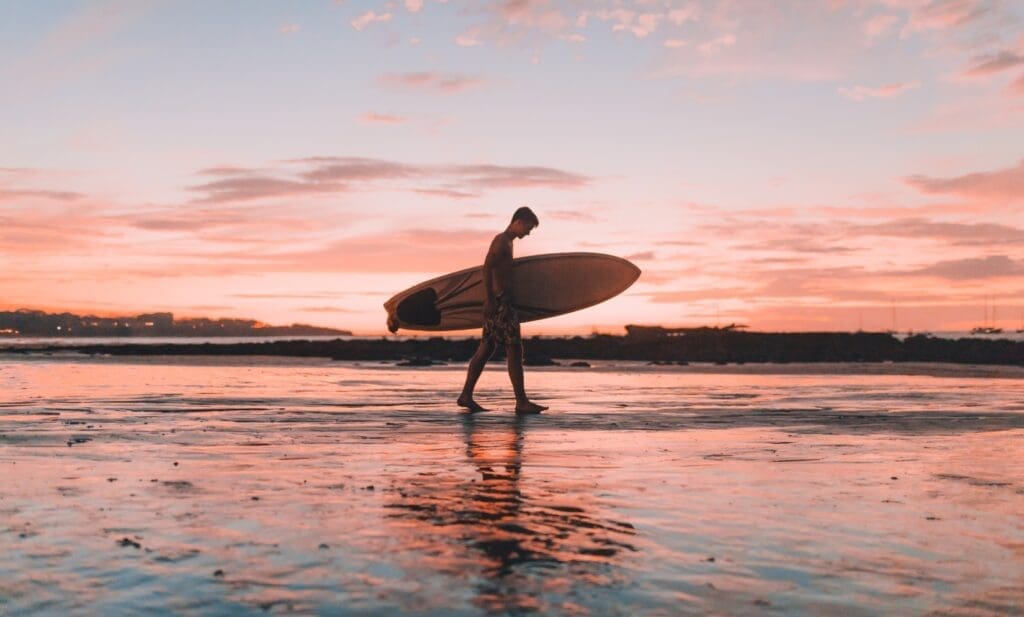 surfer holding his board while walking on the beach shoreline during sunset