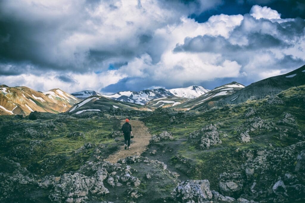 Person walking on a dirt trail in a rocky grassy valley toward snow-dusted mountains on a cloudy day