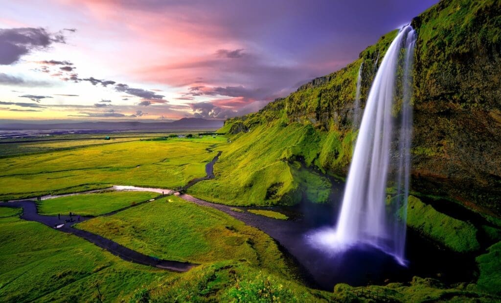 Tall waterfall cascading into a small body of water surround by lush green valley in Iceland at sunset