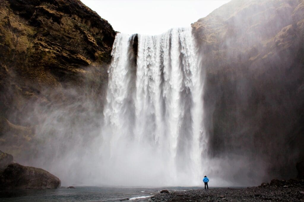 Person standing on rocky land in front of a mist created by a large waterfall crashing in a body of water