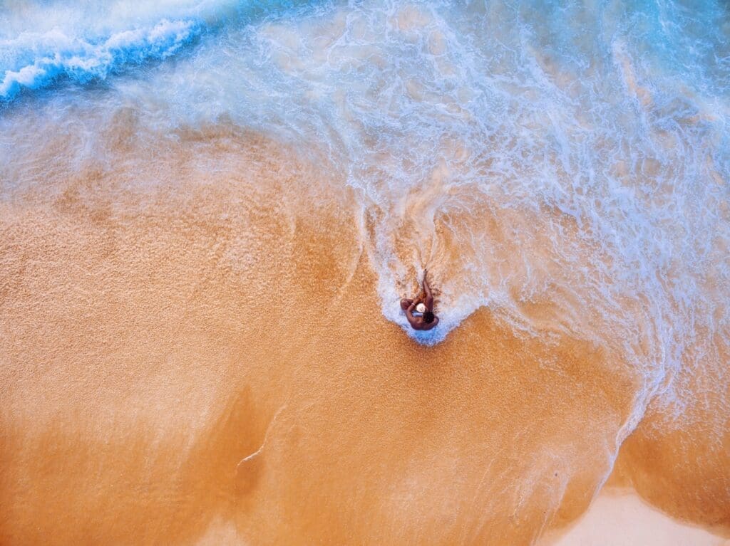 Aerial view of man sitting alone in the blue waves on a sandy beach.