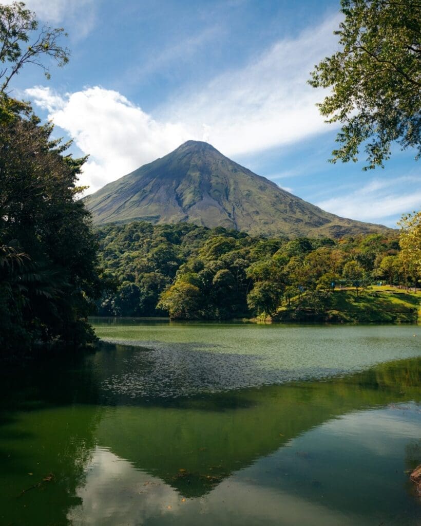 A lake sits at the forefront of the image as focus shifts to the mighty Arenal Volcano emerging from dense jungle