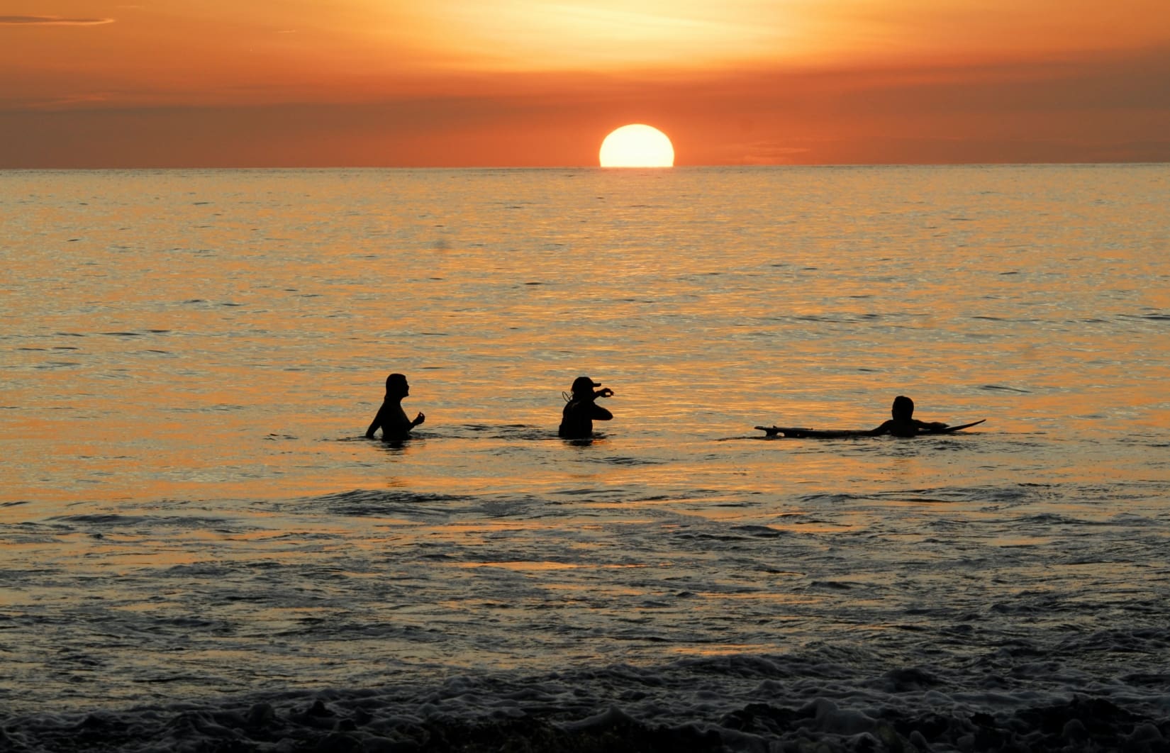 Three people swimming and surfing on the beach during a sunset